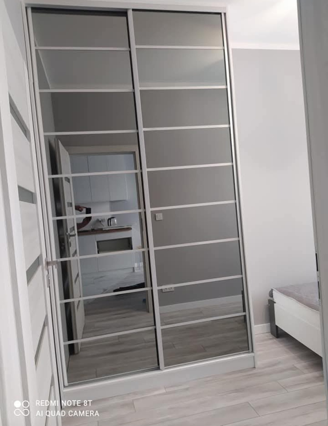 Wnękowa szafa z przesuwnymi drzwiami, z lustrzanymi panelami i białymi profilami, odbijająca wnętrze kuchni w nowoczesnym mieszkaniu.