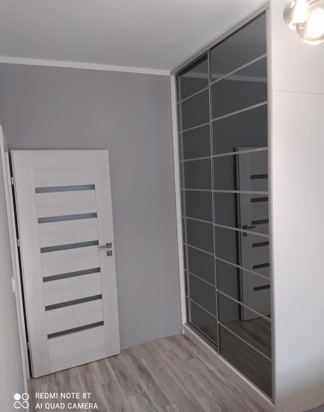 Biała szafa wnękowa z przesuwnymi drzwiami z ciemnymi szybami, widoczna na tle szarej ściany i białych drzwi z poziomymi przeszkleniami w nowoczesnym wnętrzu.