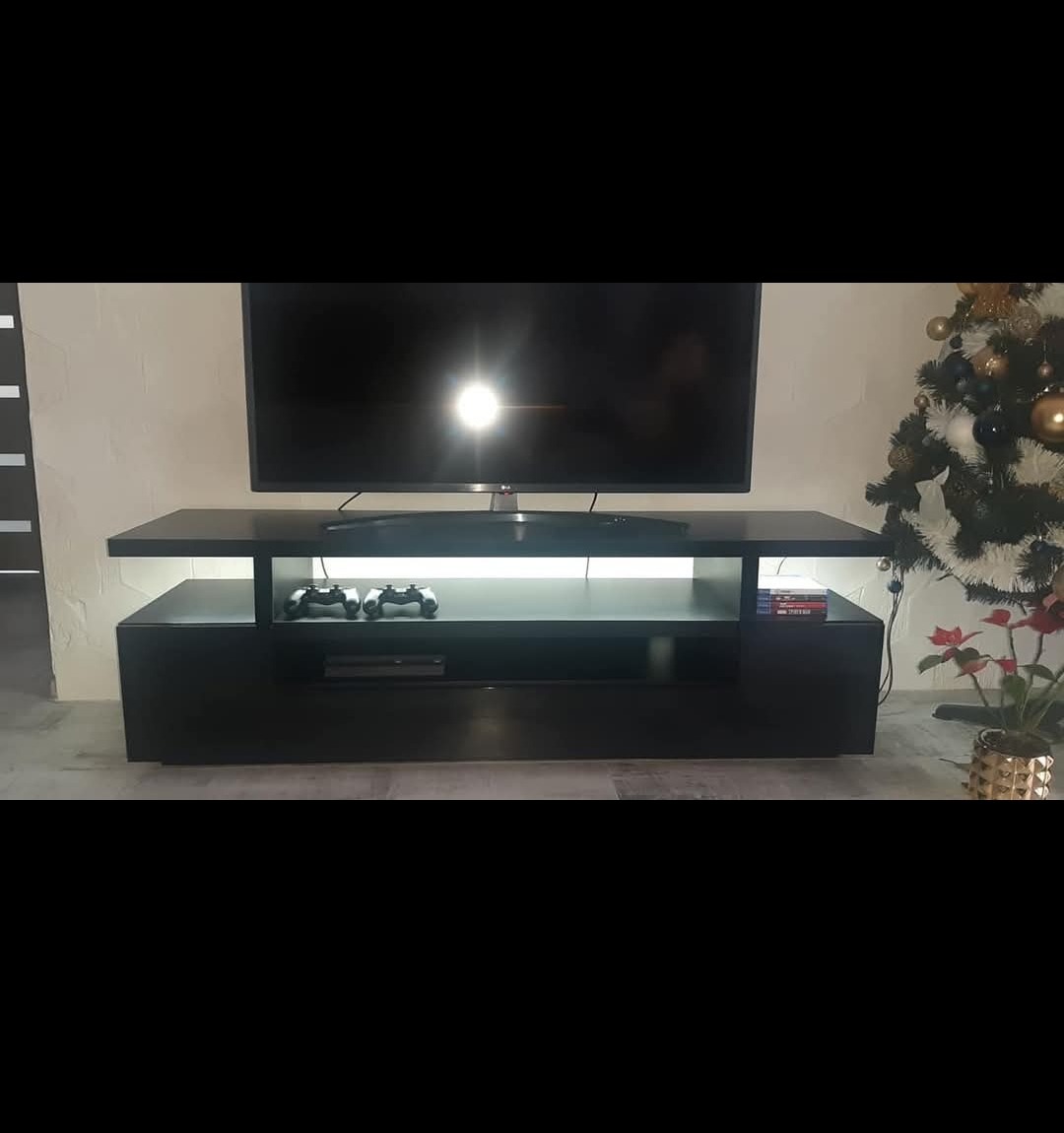 Czarna szafka RTV z podświetleniem LED, na której stoi telewizor. Na półce widoczne pady do konsoli. Obok choinka.