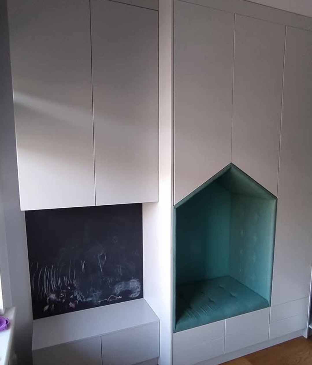 Biała szafa wnękowa z siedziskiem w kształcie domku, obok tablica kredowa. Nowoczesny design, idealne rozwiązanie do pokoju dziecięcego.
