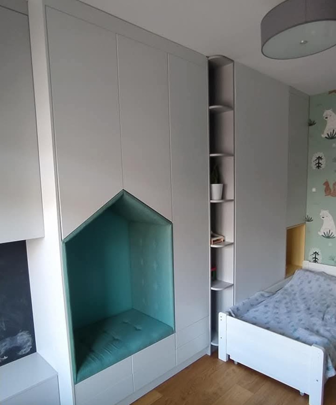 Biała szafa wnękowa z siedziskiem w kształcie domku w pokoju dziecięcym, obok regał z półkami i łóżko. Tapeta w zwierzątka w tle.
