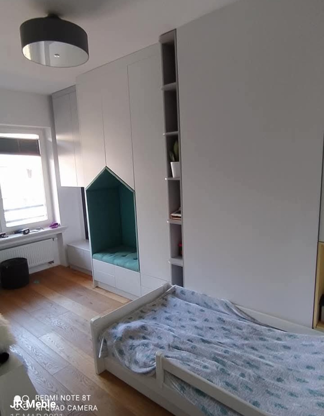 Biała szafa wnękowa z wbudowanym siedziskiem w kształcie domku i otwartymi półkami. Dziecięce łóżko na pierwszym planie. Drewniana podłoga.