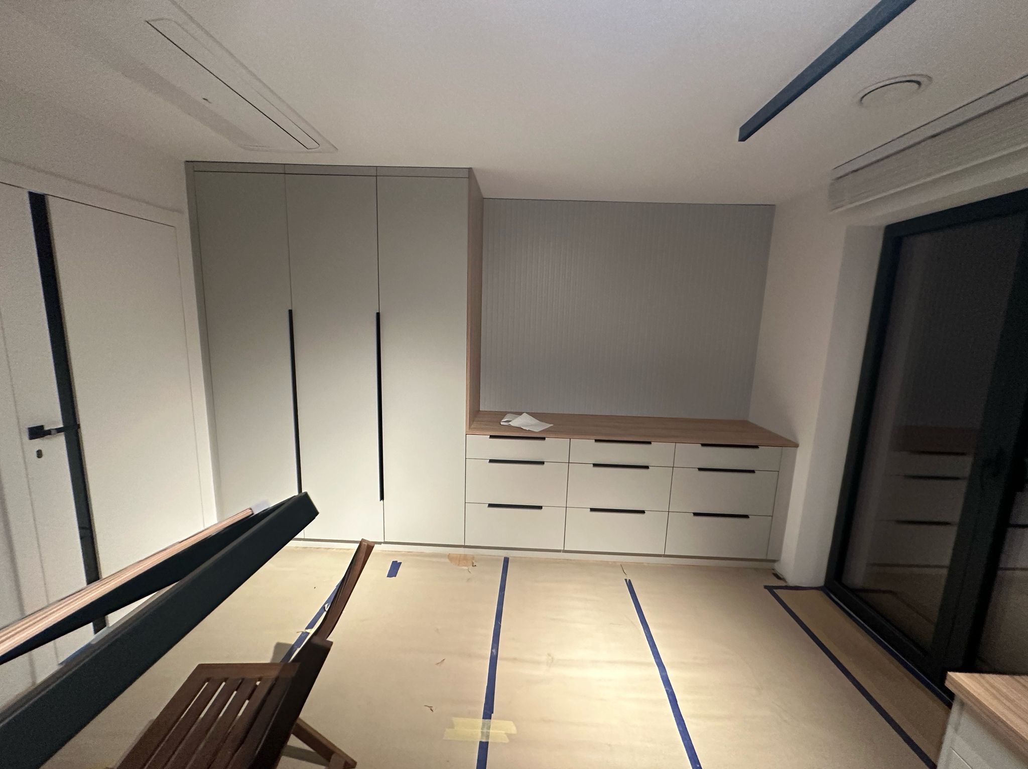 Jasne meble na wymiar: szafa z długimi uchwytami i komoda z szufladami, drewniany blat. Podłoga zabezpieczona taśmą malarską. Widok z dołu, wnętrze w trakcie wykańczania.