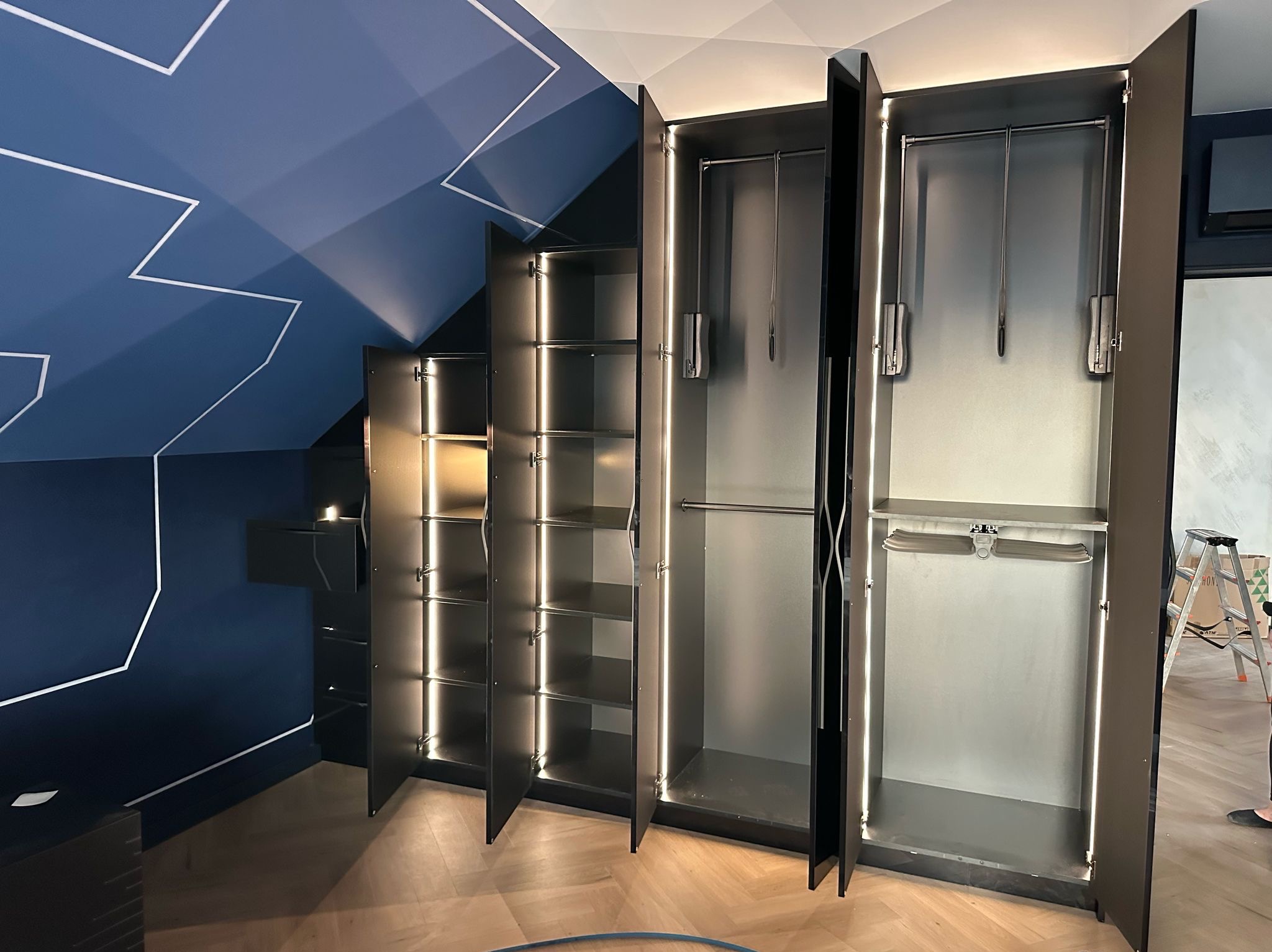 Czarna szafa na wymiar z oświetleniem LED, otwarte drzwi ukazują półki i wieszaki. Ściana w tle z geometrycznym wzorem. Widoczny fragment podłogi z parkietem.