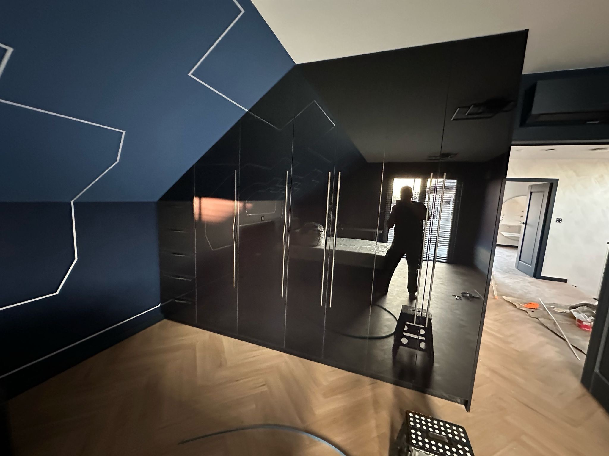 Czarna, błyszcząca szafa wnękowa na poddaszu, z widocznym odbiciem wnętrza pokoju i sylwetką osoby. Dekoracyjna linia na granatowej ścianie.