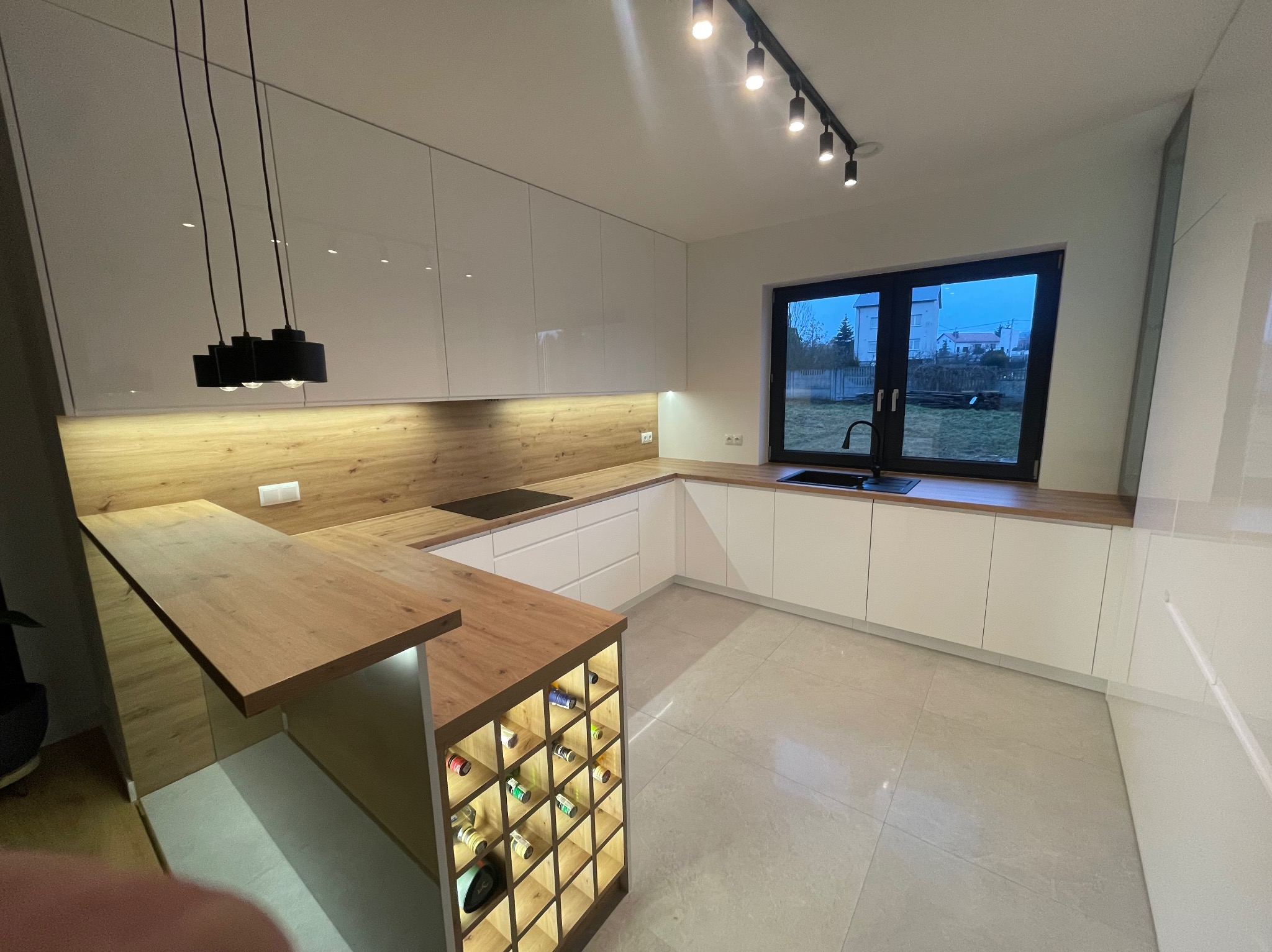 Nowoczesna, jasna kuchnia z białymi frontami, drewnianym blatem i wyspą z podświetlanym barkiem na wino. Nad blatem wiszą czarne lampy.