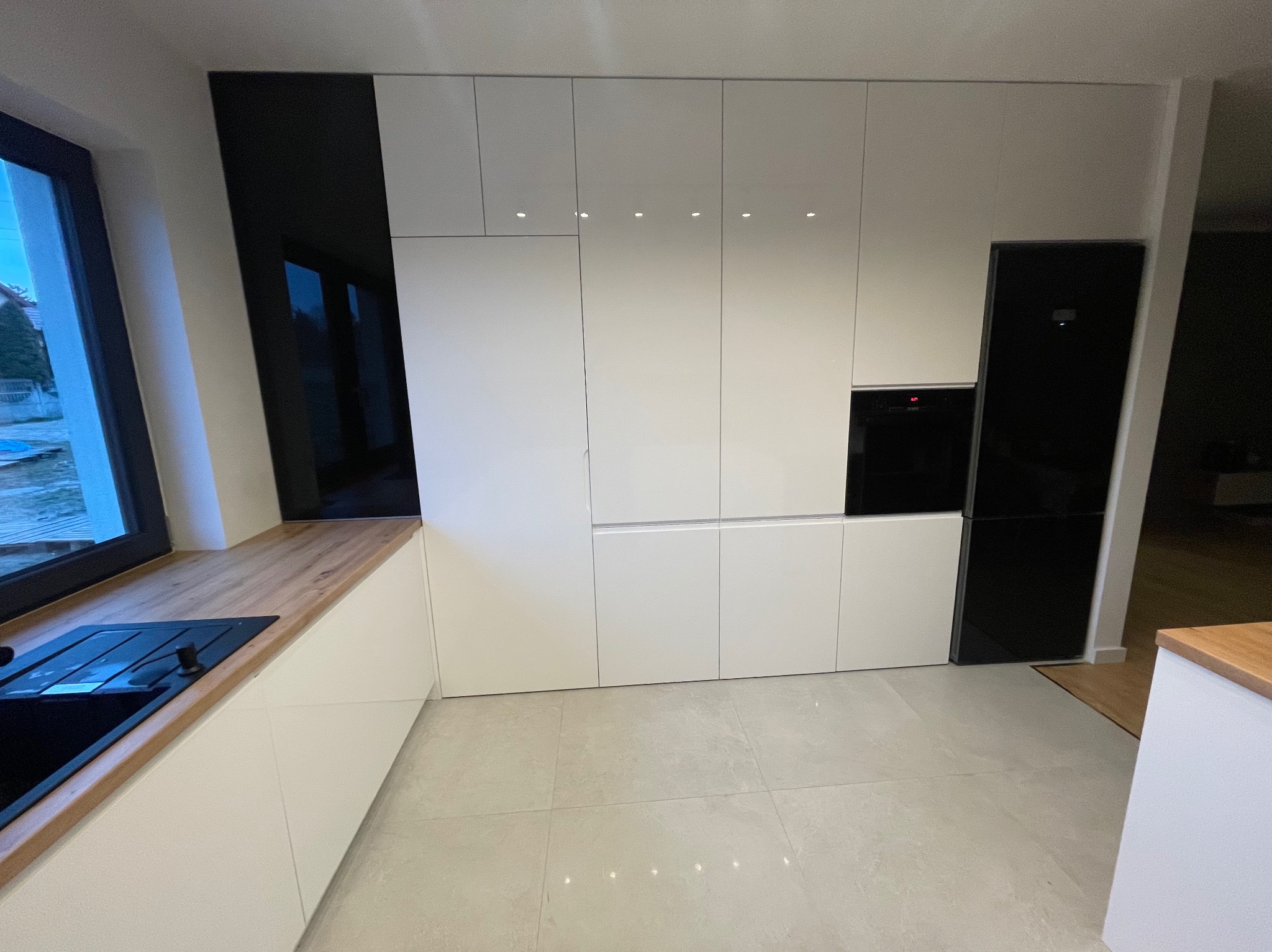 Nowoczesna, minimalistyczna kuchnia z białymi frontami i czarnymi akcentami. Widoczny blat z drewnianym wykończeniem i czarny zlew. Całość dopełnia jasna podłoga.