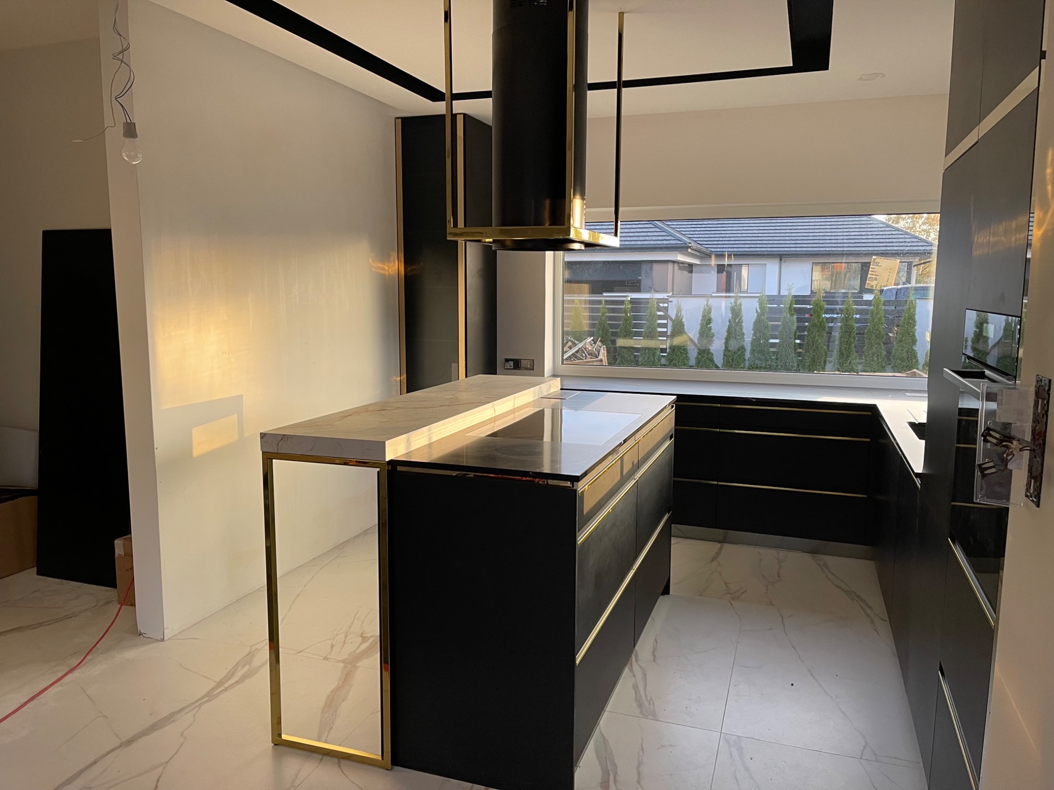 Nowoczesna, czarno-złota kuchnia z wyspą i marmurowym blatem, widok na okno z zielenią. Minimalistyczny design, surowy stan wykończenia pomieszczenia.