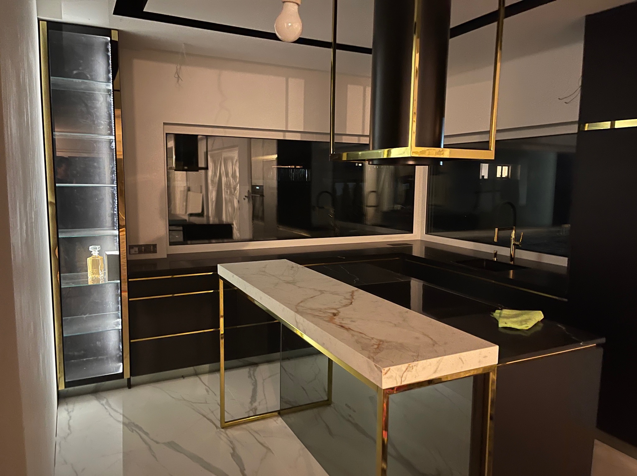 Nowoczesna kuchnia z czarnymi szafkami, złotymi akcentami i marmurowym blatem wyspy. Widoczna witryna z oświetleniem i dekoracyjnym flakonem.