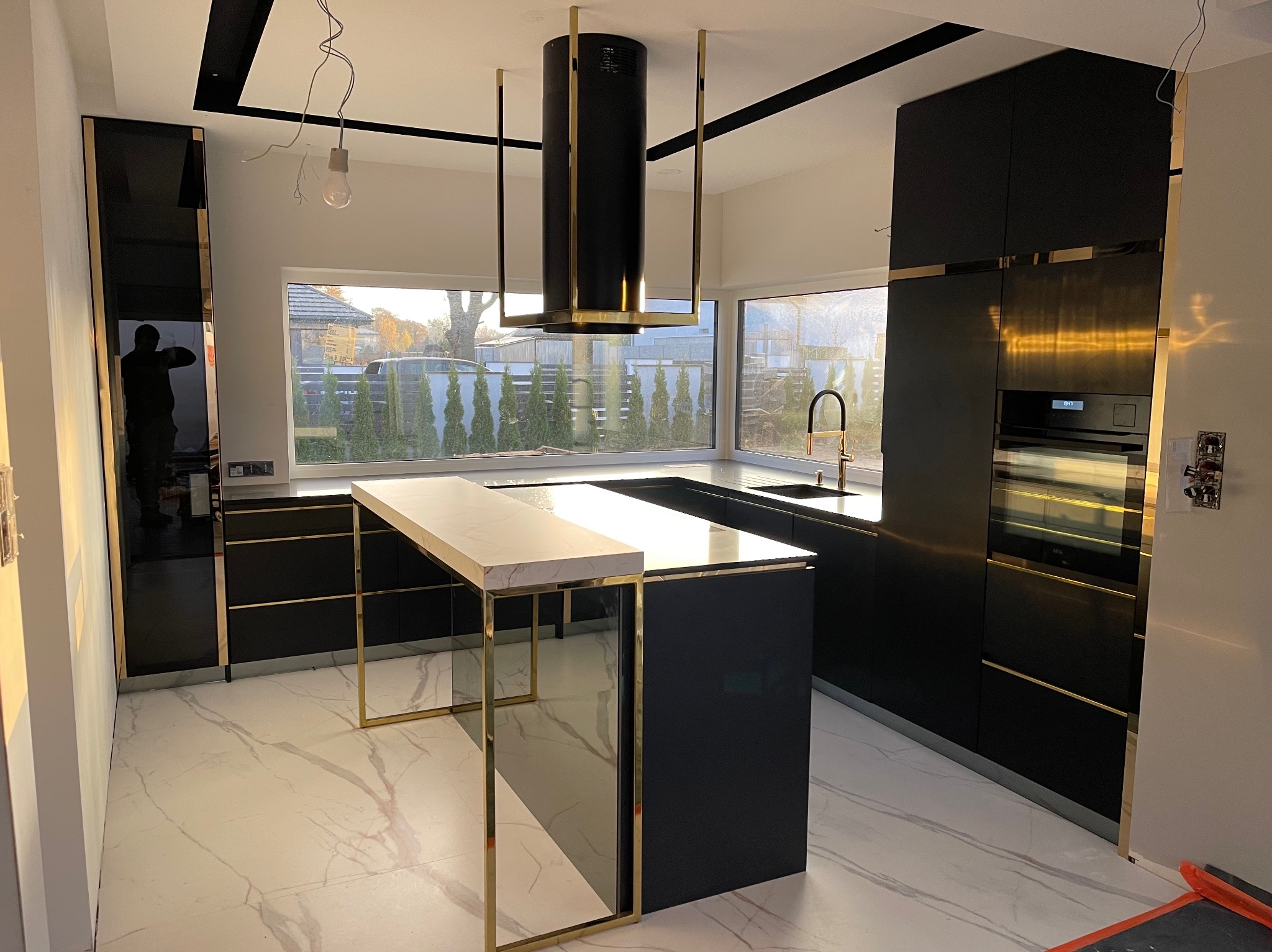 Nowoczesna, czarna kuchnia z wyspą i złotymi akcentami. Minimalistyczny okap cylindryczny. Marmurowa podłoga, duże okno z widokiem na zieleń.
