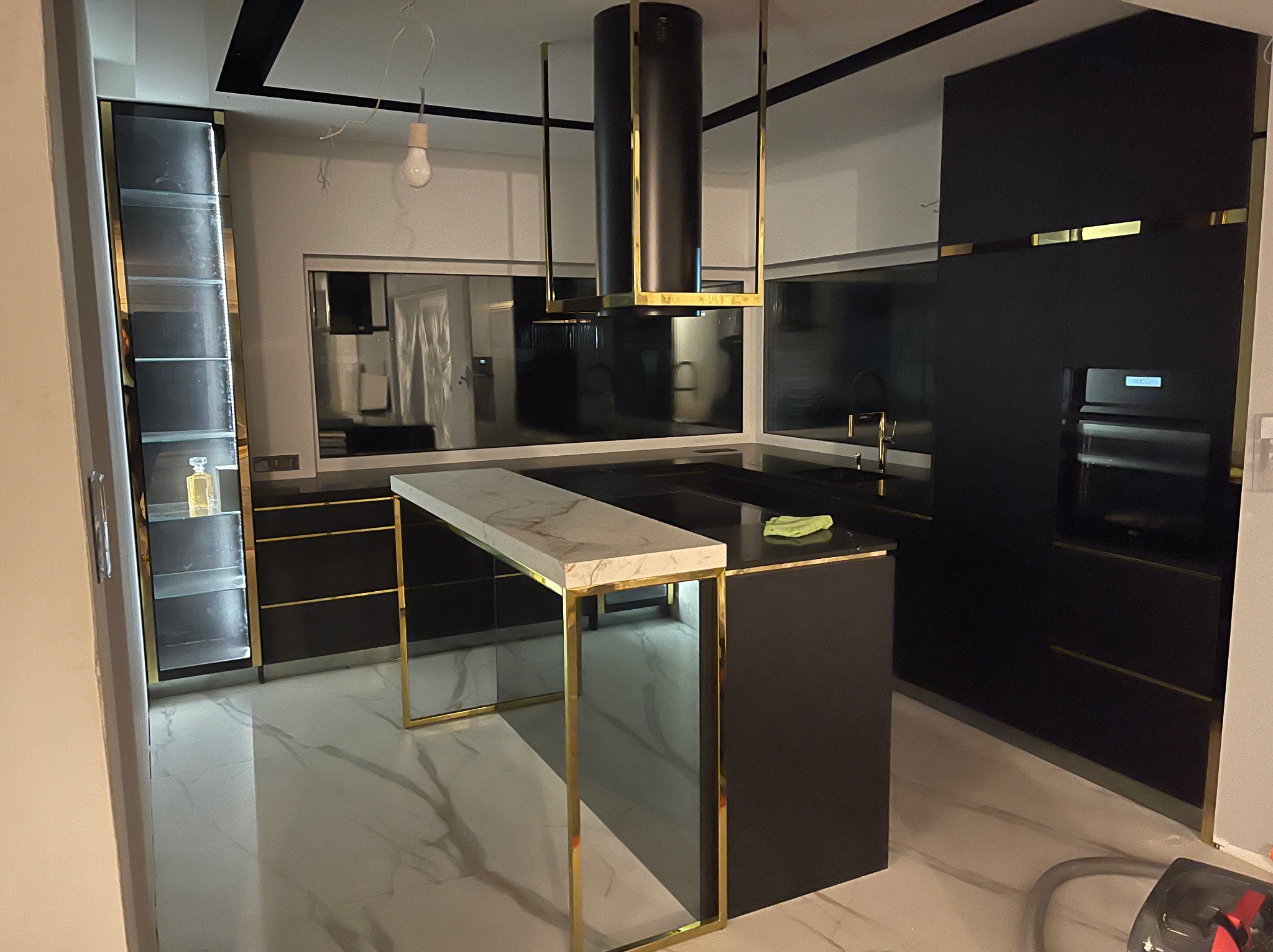 Nowoczesna, czarna kuchnia z wyspą o marmurowym blacie i złotych detalach. Uwagę przykuwa designerski okap i przeszklona witryna z oświetleniem.