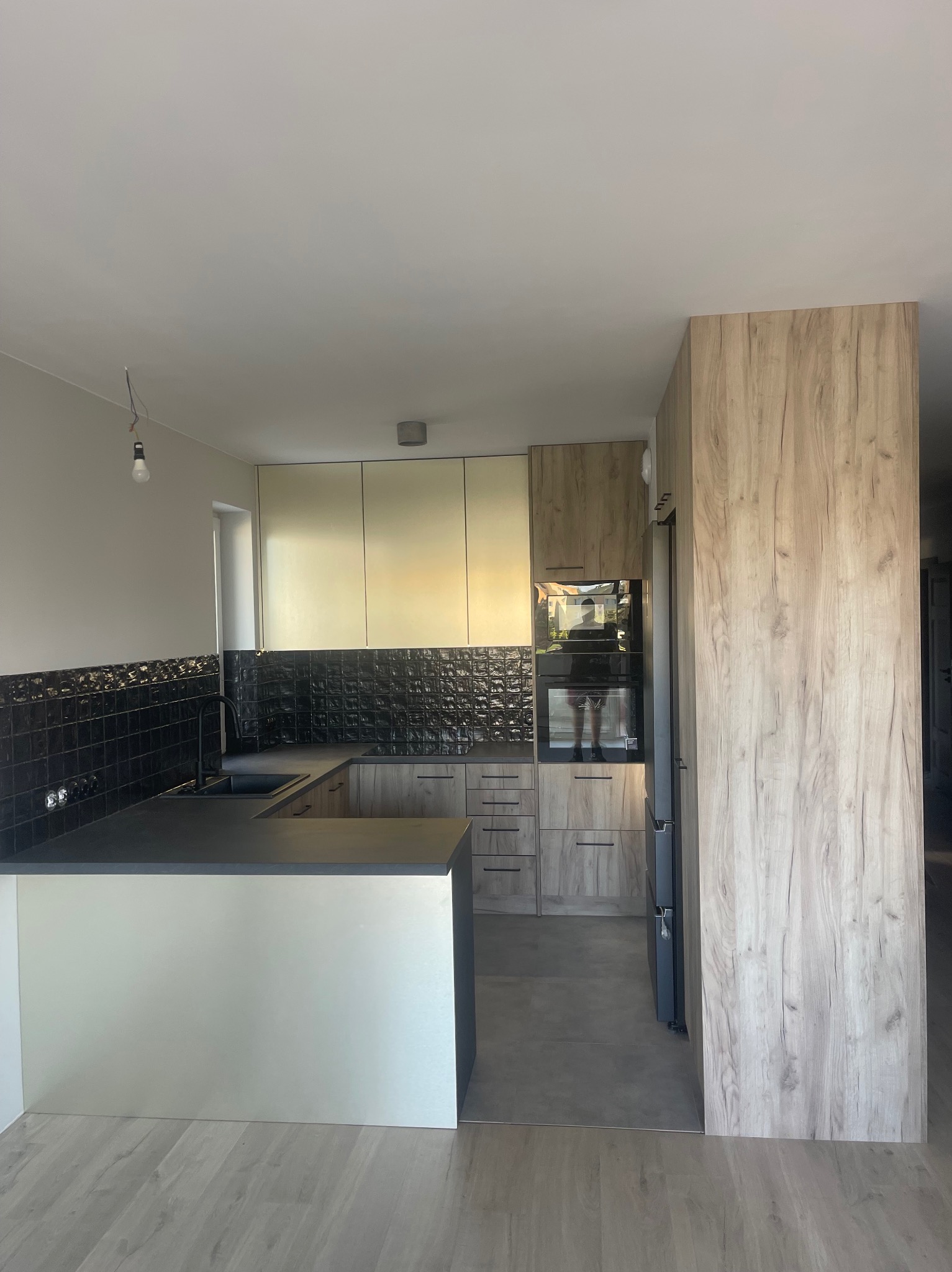 Nowoczesna kuchnia z wyspą i czarnymi płytkami. Jasne fronty szafek górnych kontrastują z drewnopodobnymi dolnymi. Widoczny piekarnik i lodówka.