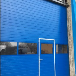 Niebieska brama garażowa segmentowa z oknami i drzwiami wejściowymi, widok z zewnątrz. Metalowa konstrukcja nośna. Świeżo zamontowana.