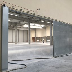 PROLINE PIOTR BURAKOWSKI - Otwarta, przesuwna brama przemysłowa w hali, z widoczną instalacją przeciwpożarową i osobą w oddali. Brama metalowa, szara, z widocznymi elementami mocującymi.