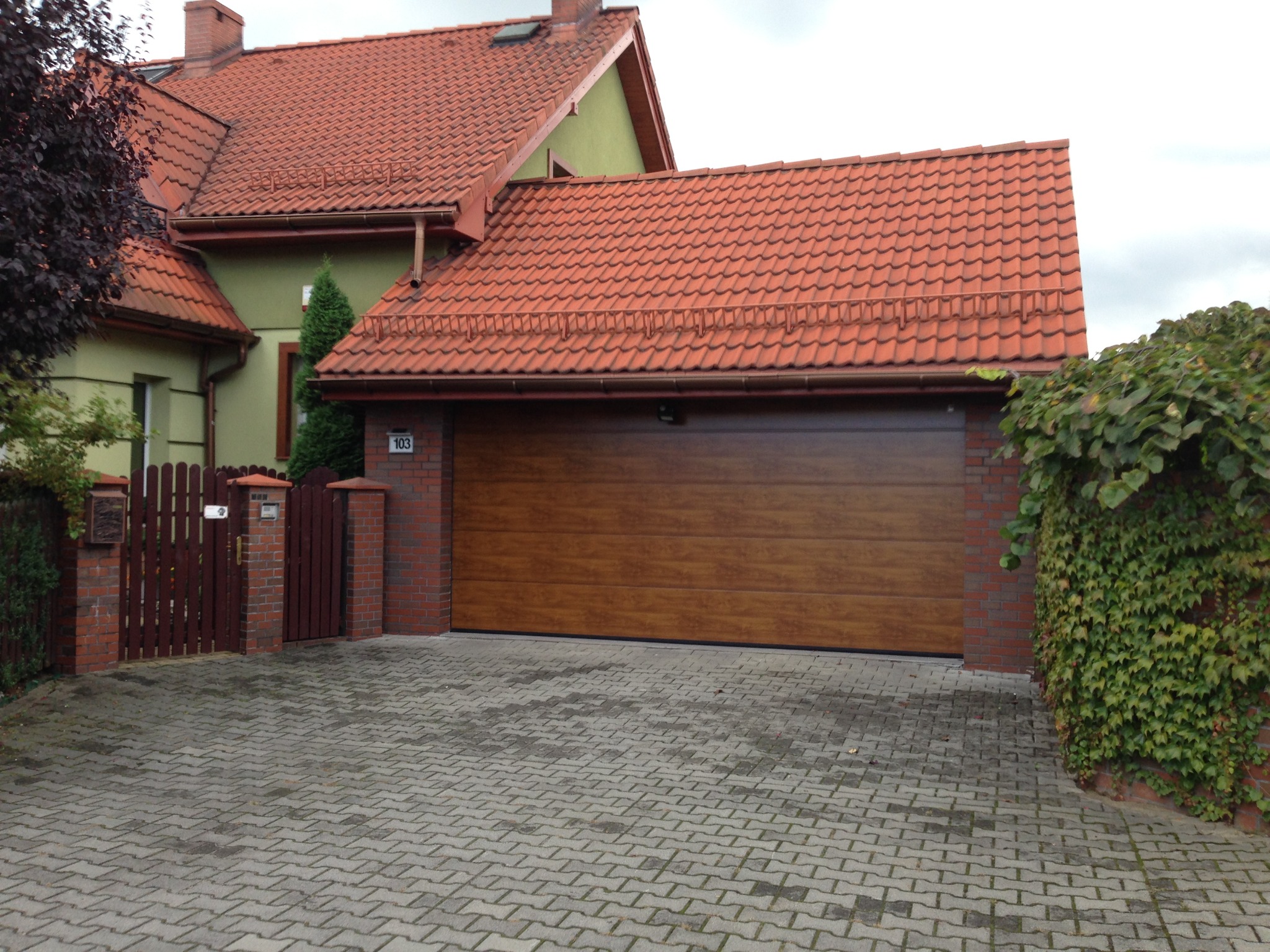 Nowoczesna, segmentowa brama garażowa w kolorze drewna, wkomponowana w bryłę domu z czerwoną dachówką i ceglanym ogrodzeniem. Kostka brukowa na podjeździe.