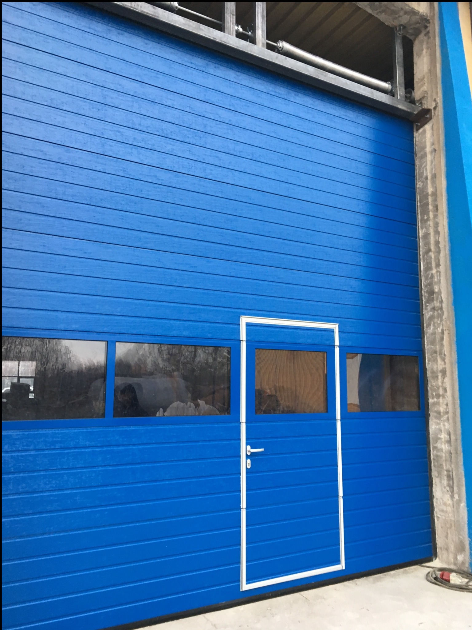 Niebieska brama garażowa segmentowa z oknami i drzwiami wejściowymi, widok z zewnątrz. Metalowa konstrukcja nośna. Świeżo zamontowana.