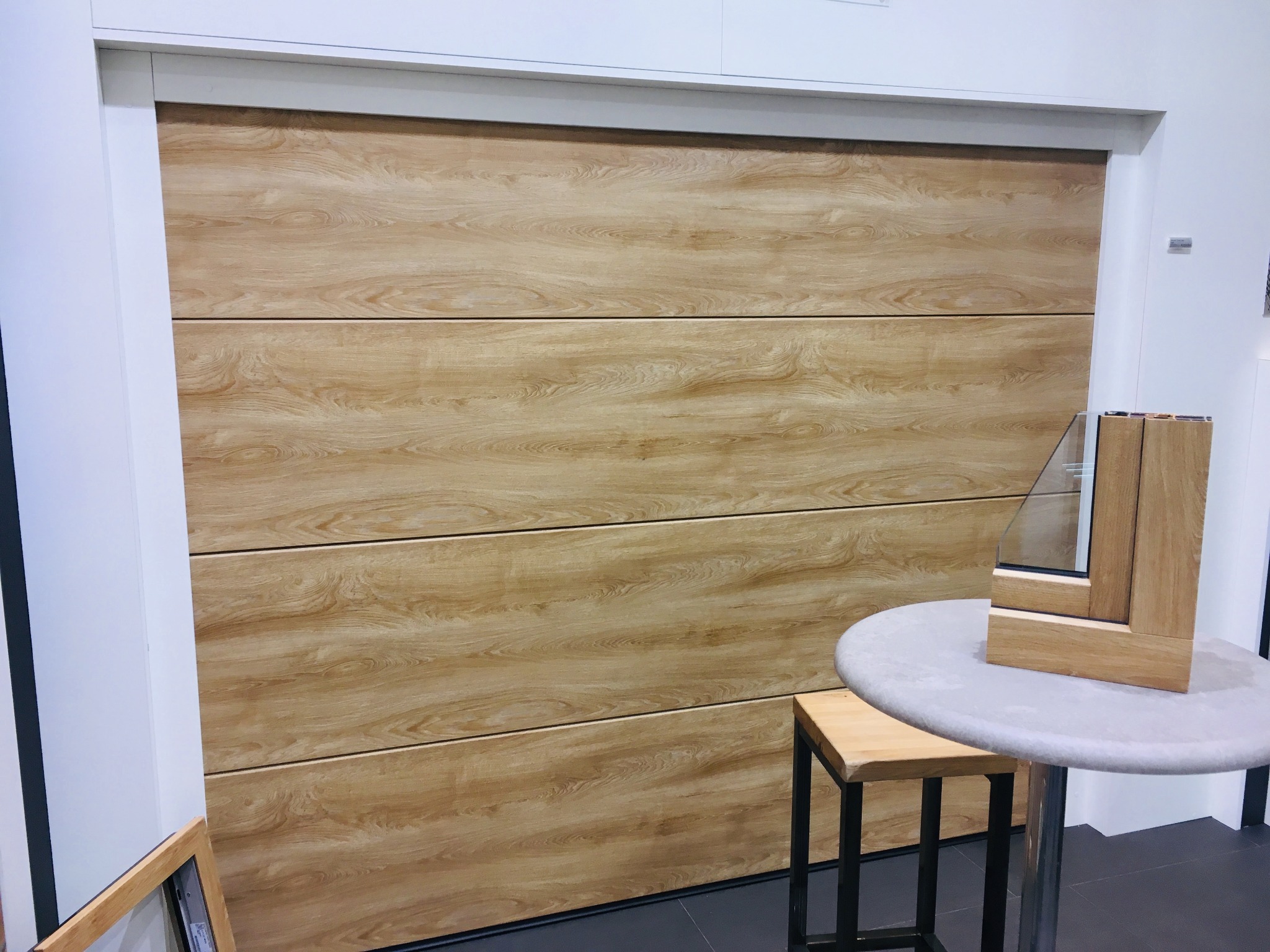 Drewnopodobna brama garażowa segmentowa z poziomym wzorem paneli, prezentowana obok przekroju okna na stole. Brama w jasnej kolorystyce drewna.