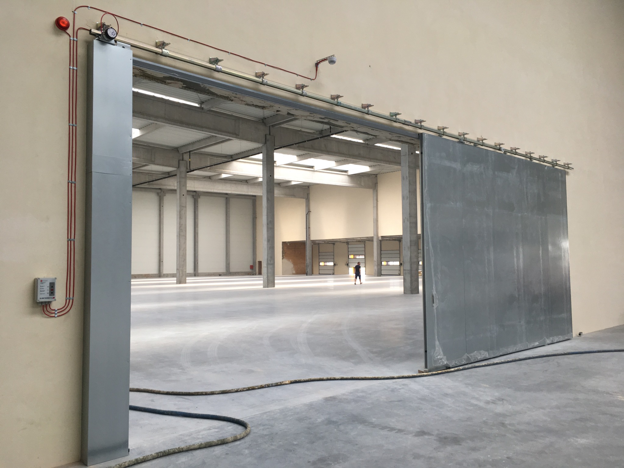 Otwarta, przesuwna brama przemysłowa w hali, z widoczną instalacją przeciwpożarową i osobą w oddali. Brama metalowa, szara, z widocznymi elementami mocującymi.