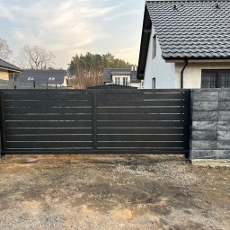 SteelCraft Andrii Hodziur - Nowoczesna, przesuwna brama wjazdowa z poziomych profili, zamontowana na betonowych słupkach. Tło stanowią domy jednorodzinne i drzewa.