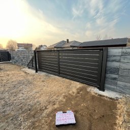 SteelCraft Andrii Hodziur - Nowoczesna, przesuwna brama wjazdowa z poziomymi panelami, zamontowana na tle szarego, betonowego muru. Na pierwszym planie skrzynka narzędziowa i przygotowany teren.