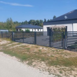 Usługi ogólnobudowlane Robert Chodkowski - Nowoczesne, poziome ogrodzenie metalowe w kolorze grafitowym, z bramą wjazdową, na tle domu jednorodzinnego i błękitnego nieba. Widok z zewnątrz posesji.