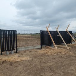Usługi ogólnobudowlane Robert Chodkowski - Częściowo zamontowana, czarna, metalowa brama przesuwna z drewnianymi podporami na tle ogrodzenia panelowego i pochmurnego nieba. Widok z zewnątrz posesji.