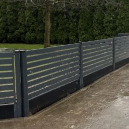 Usługi ogólnobudowlane Robert Chodkowski - Nowoczesne, poziome ogrodzenie panelowe w kolorze grafitowym, z widocznymi słupkami i podmurówką. W tle zielone tuje i drzewo bez liści. Brukowana nawierzchnia.