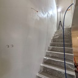 Aliaksej Kruty - Świeżo pomalowana, jasna ściana przy betonowych schodach w trakcie wykańczania. Widoczne puszki elektryczne i kabel. Scena oświetlona lampą budowlaną.