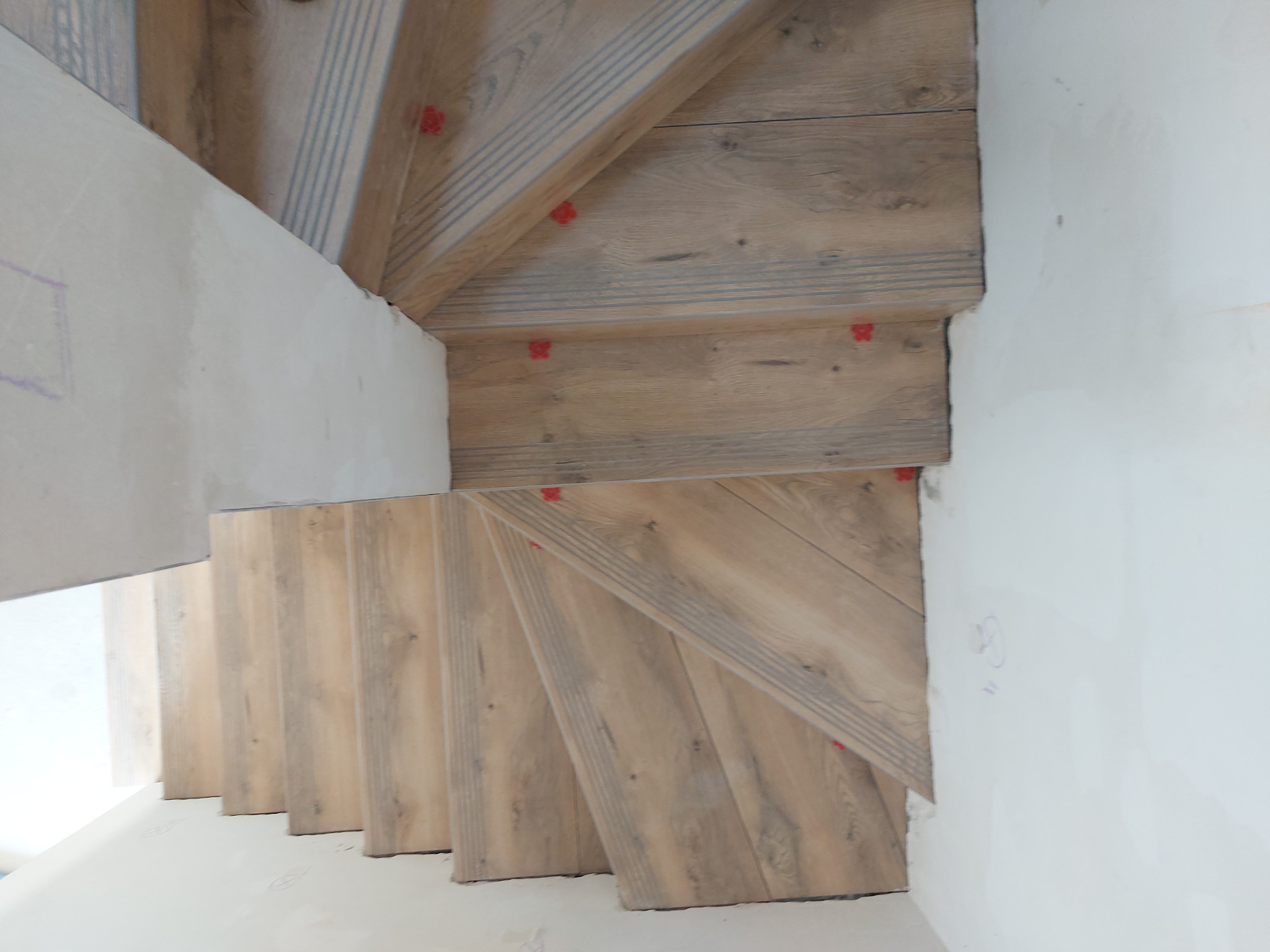 Ułożone płytki imitujące drewno na schodach spiralnych, z czerwonymi elementami poziomowania. Ściany w stanie surowym, widoczne szkice. Ujęcie z góry.