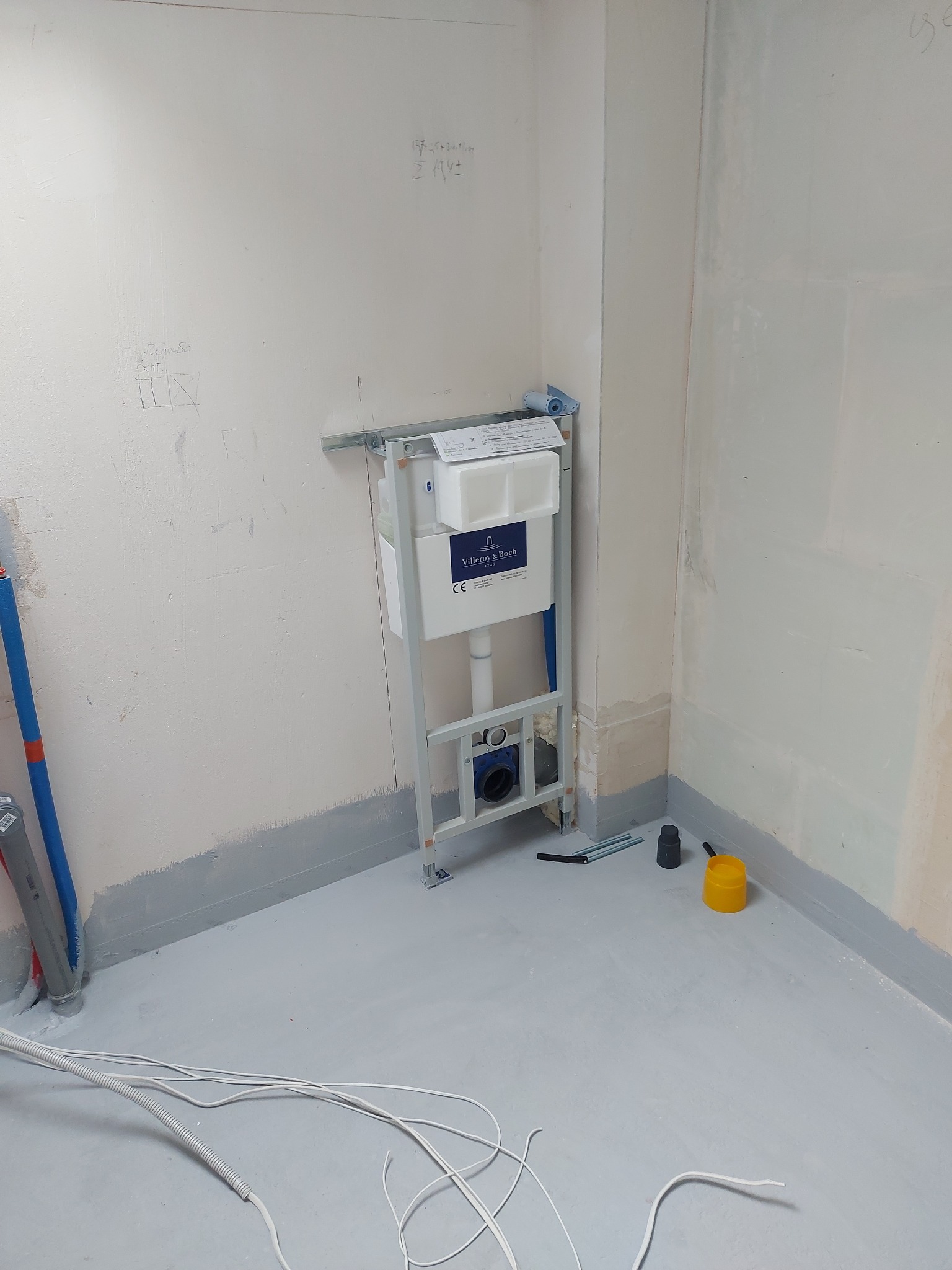 Stelaż podtynkowy Villeroy&Boch zamontowany na ścianie w trakcie remontu łazienki. Szare ściany z widocznymi śladami po pracach wykończeniowych. Na podłodze kable.