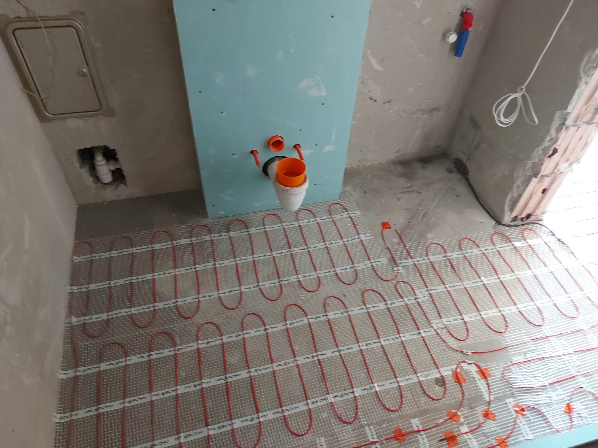 Instalacja ogrzewania podłogowego w łazience przed wylewką: czerwone kable grzewcze ułożone na siatce, pion kanalizacyjny przy ścianie z płyt gipsowych.