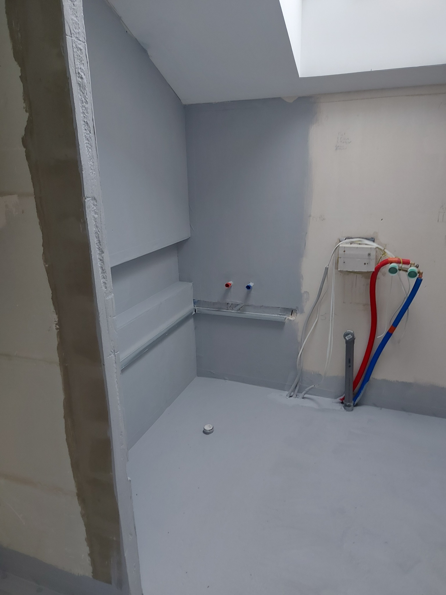 Szaro-białe wnętrze łazienki w trakcie prac wykończeniowych. Widoczne rury, surowy tynk i pomalowane ściany. Oświetlenie górne.