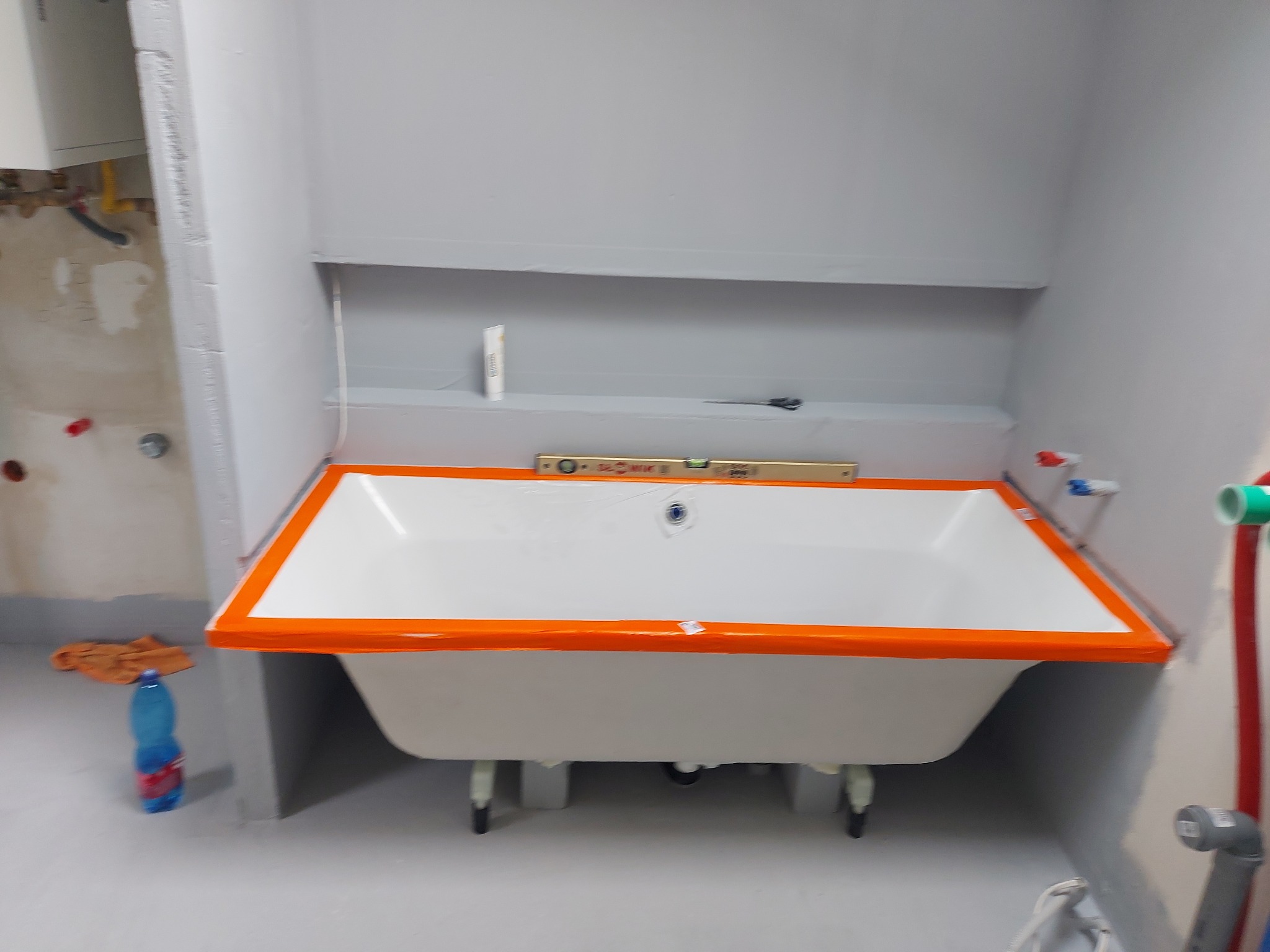 Biała wanna z pomarańczowym zabezpieczeniem krawędzi, poziomica na wannie, szare ściany w trakcie malowania, niebieska butelka wody na podłodze.