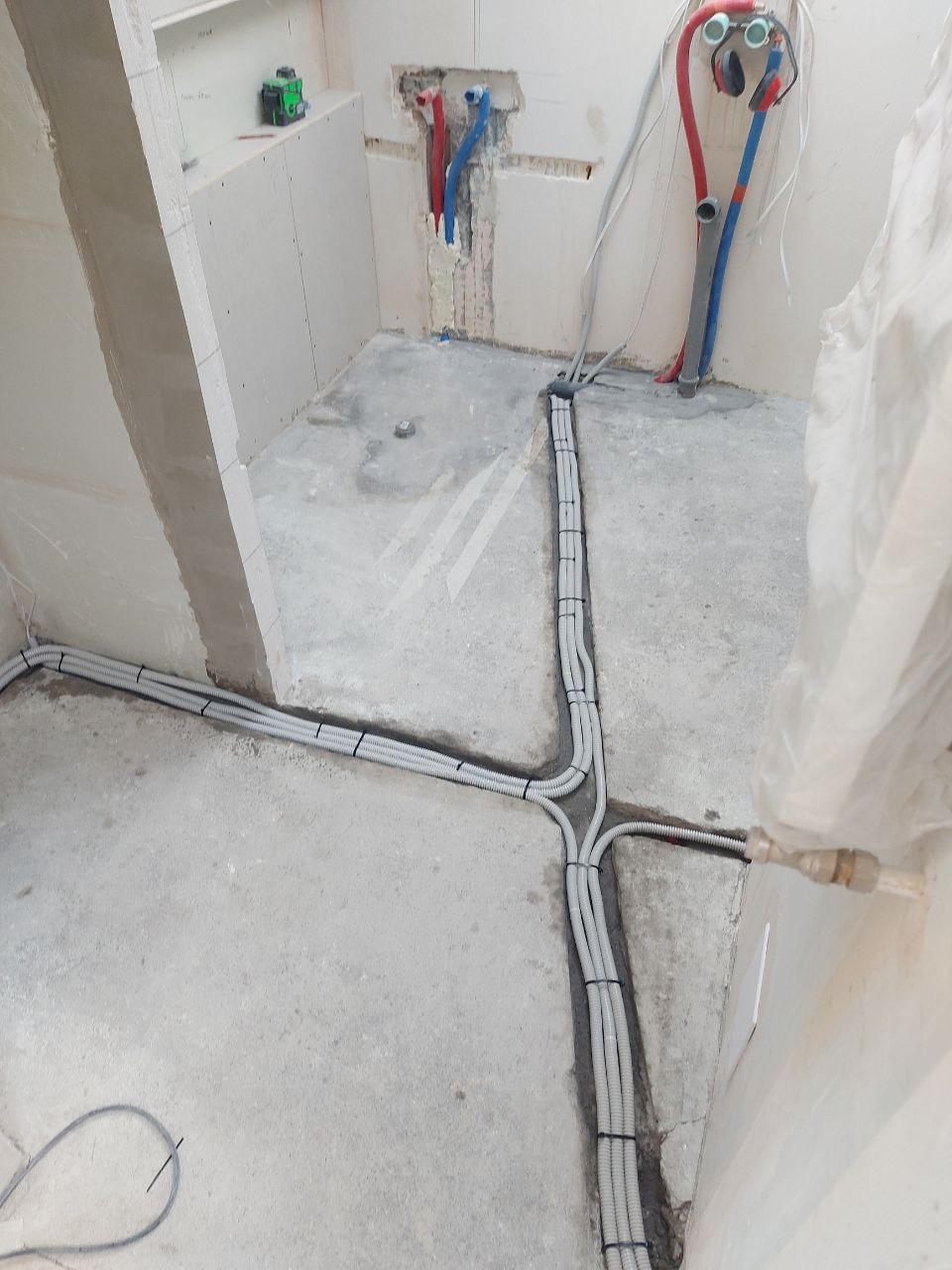 Szare rury instalacji elektrycznej ułożone w bruzdach na podłodze w surowym wnętrzu. Widoczne piony wod-kan i laser krzyżowy na półce z płyt g-k.