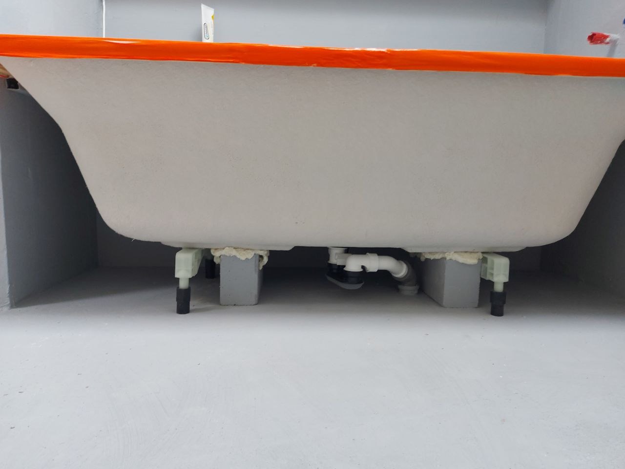 Szara wanna z pomarańczowym obramowaniem, wsparta na blokach, z widoczną instalacją hydrauliczną, na tle szarej ściany i podłogi. Detal techniczny po montażu.