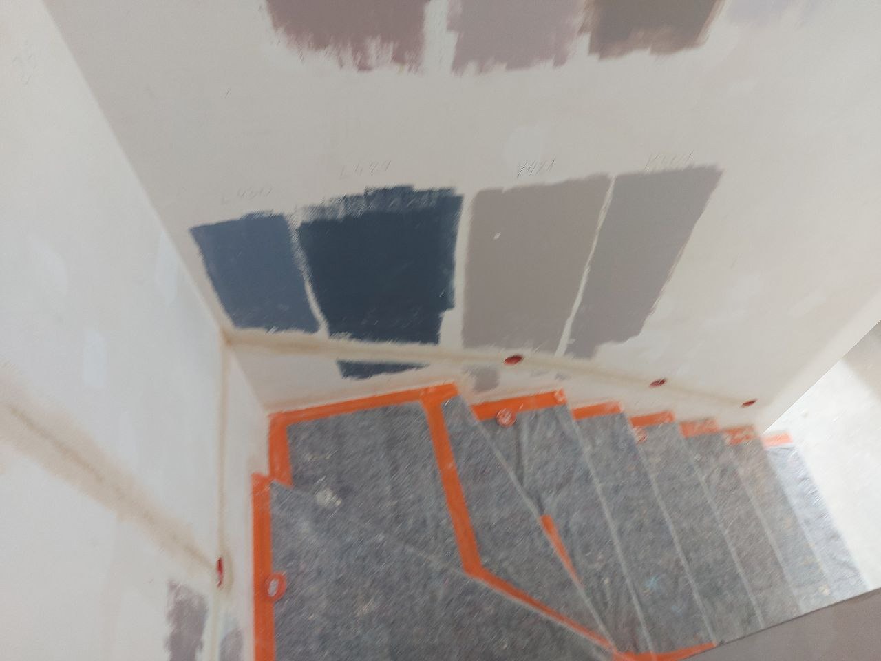 Ściana z próbkami farb w różnych odcieniach szarości nad zabezpieczonymi schodami. Testowanie kolorów przed malowaniem w mieszkaniu.