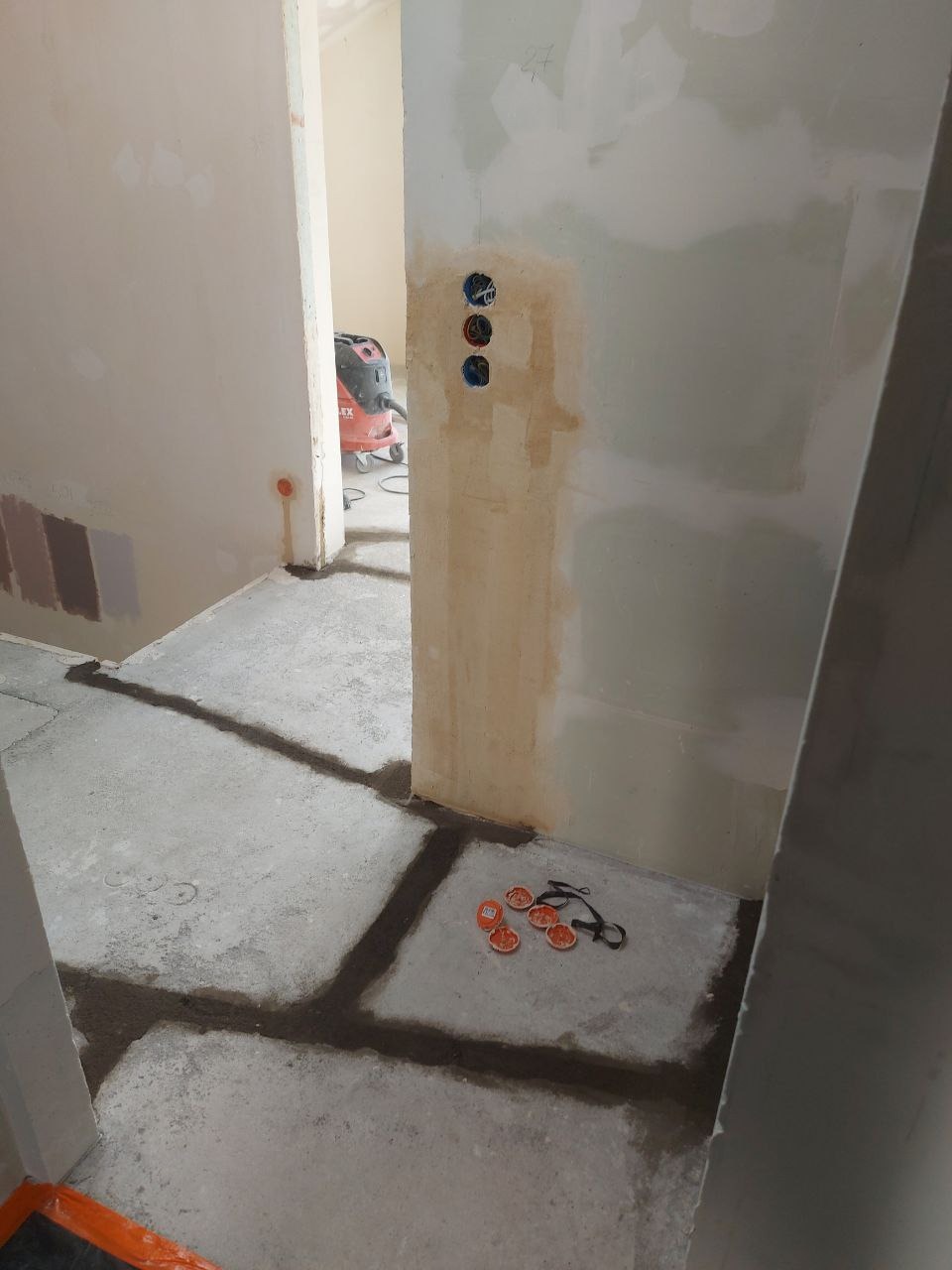 Przygotowanie do malowania: ściany z gładzią gipsową, zabezpieczona podłoga, widoczne puszki elektryczne i odkurzacz przemysłowy w tle. Etap prac wykończeniowych.