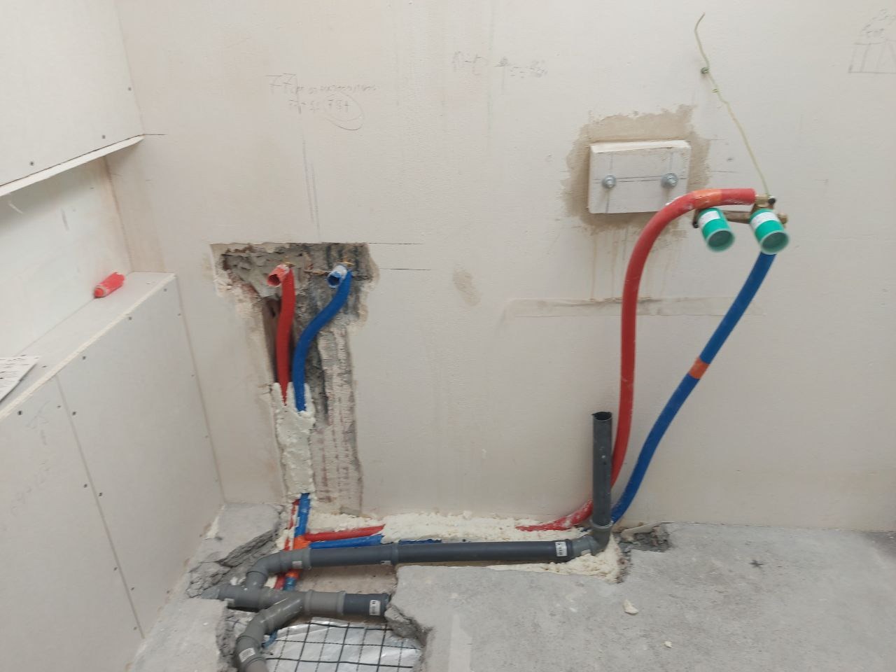 Instalacja wodno-kanalizacyjna w trakcie montażu: rury czerwone i niebieskie wystają ze ściany, szare rury na podłodze, widoczna siatka wzmacniająca w podłodze.
