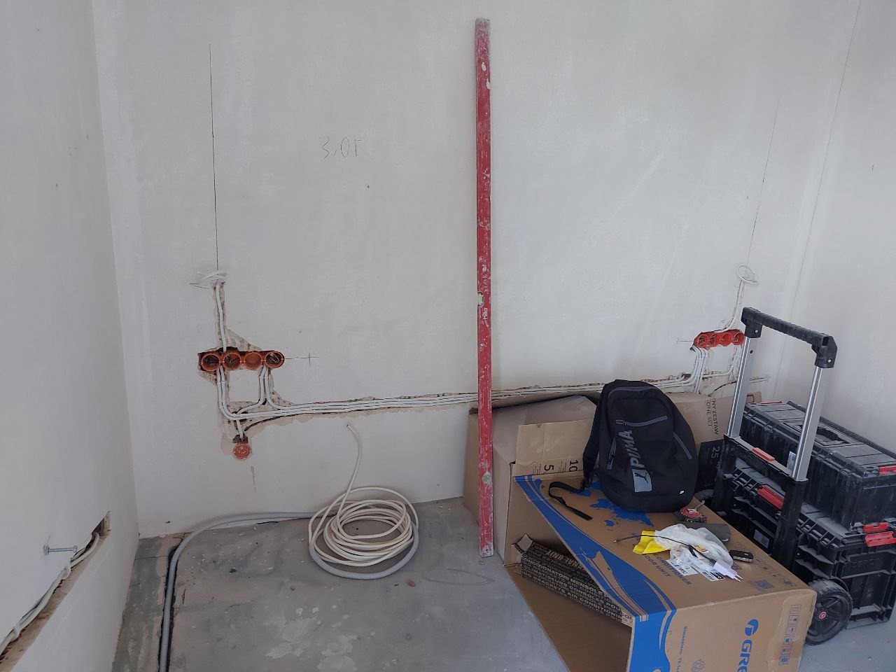 Stan surowy mieszkania w trakcie remontu: widoczne kable elektryczne w ścianie, poziomica, kartony i narzędzia. Przygotowanie do prac wykończeniowych.