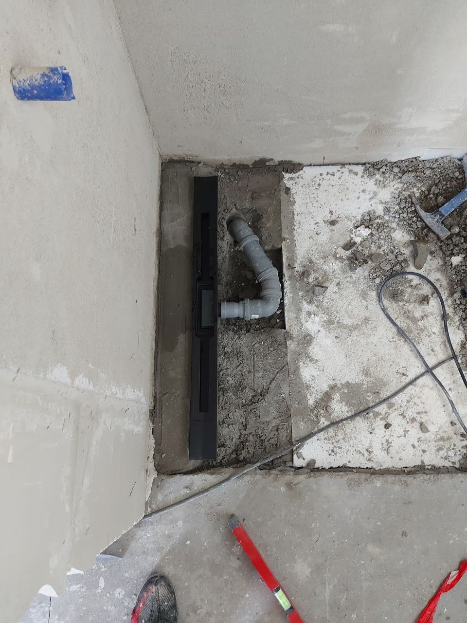 Montaż odpływu liniowego w łazience podczas remontu. Widoczne rury kanalizacyjne, poziomica i fragment buta montera. Surowy stan, przed wykończeniem.