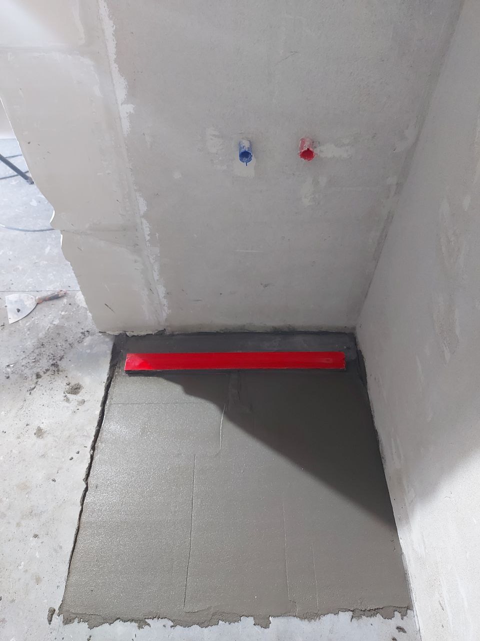 Wylewka podłogowa w łazience w trakcie prac, poziomowana czerwoną łatą. Widoczne wystające rury z instalacji wodnej.