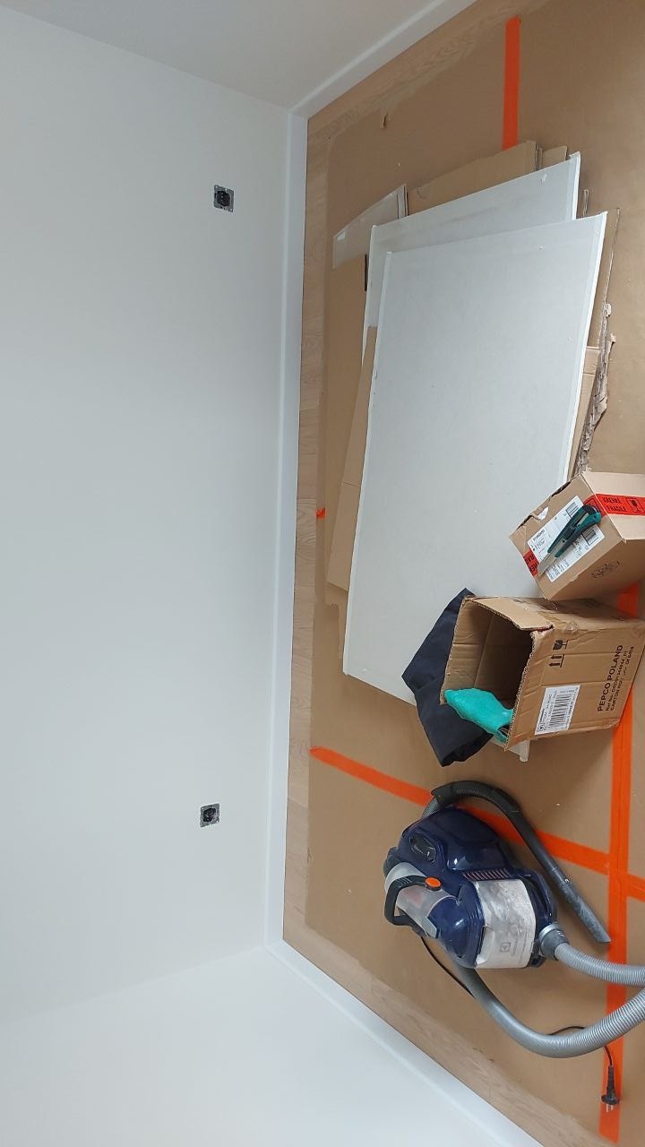 Biała ściana po malowaniu, widoczne puszki elektryczne, zabezpieczona podłoga kartonem, odkurzacz i akcesoria malarskie w kartonach.