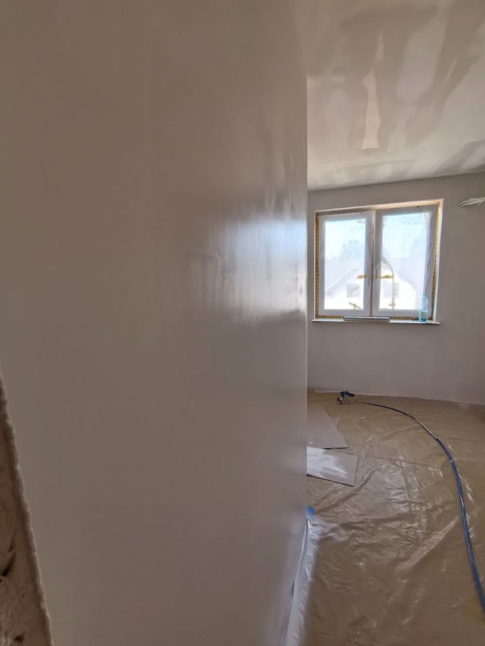 Świeżo pomalowana, gładka ściana w jasnym odcieniu, widoczna z bliska. Na podłodze folia ochronna i sprzęt malarski w tle. Okno z widokiem na zewnątrz.