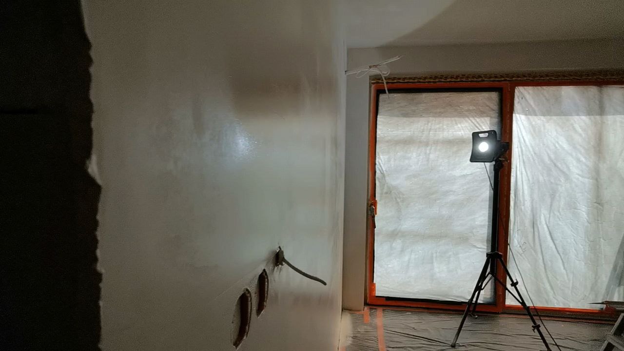 Świeżo pomalowana, jasna ściana z widocznymi puszkami elektrycznymi bez osprzętu. Okno zabezpieczone folią malarską, oświetlone lampą na statywie.