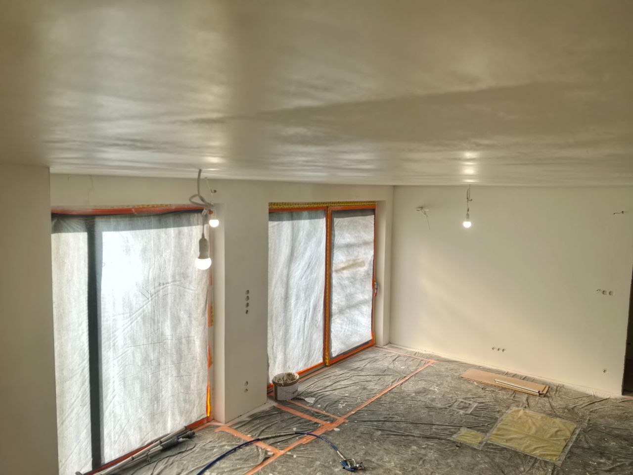 Świeżo pomalowane wnętrze pokoju z zabezpieczonymi oknami folią malarską. Widoczne narzędzia malarskie i wiaderko z farbą. Świeżo pomalowany sufit.