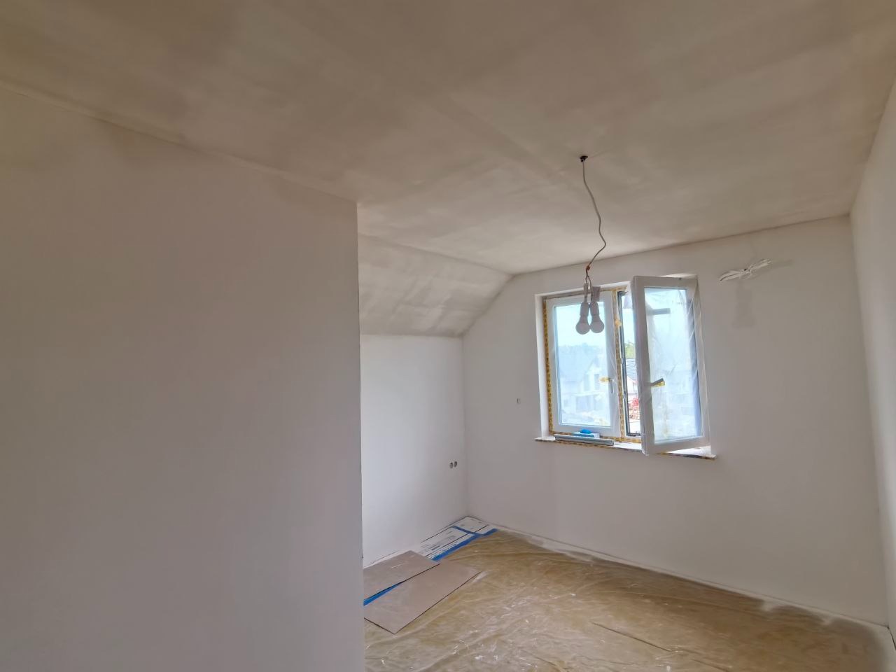 Pokój po malowaniu: białe ściany, zabezpieczone okno folią malarską, podłoga zakryta kartonem i folią. Widoczne gniazdka elektryczne. Wnętrze gotowe do użytku.