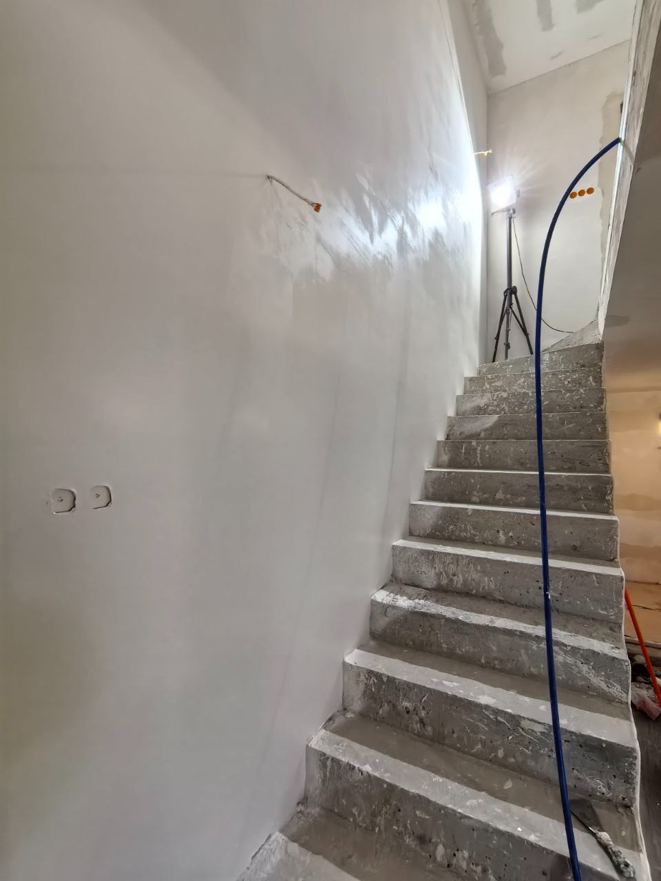 Świeżo pomalowana, jasna ściana przy betonowych schodach w trakcie wykańczania. Widoczne puszki elektryczne i kabel. Scena oświetlona lampą budowlaną.