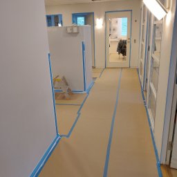 USŁUGI BUDOWLANE Krzysztof Gołębiowski - Przygotowanie do malowania: zabezpieczone taśmą i papierem ściany, podłoga w korytarzu biurowym. Widoczna drabina i białe ściany.