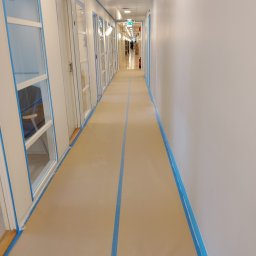 USŁUGI BUDOWLANE Krzysztof Gołębiowski - Korytarz zabezpieczony taśmą malarską przed pracami wykończeniowymi. Widoczne białe ściany i podłoga w kolorze beżowym. W tle osoba przechodząca korytarzem.