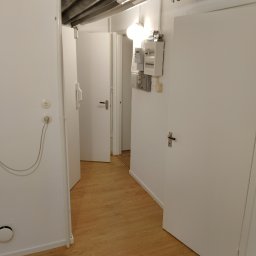 USŁUGI BUDOWLANE Krzysztof Gołębiowski - Białe, odświeżone wnętrze korytarza z drewnianą podłogą, widoczne drzwi i instalacje pod sufitem. Minimalistyczny design z elementami technicznymi.